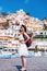 A girl in a sunglasses and a hat stands on the beach in Positano. View of houses and hotels in the background. answers the phone
