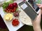 A girl takes pictures of a pizza on a smartphone. Necessary ingredients are on the table: cheese, sausage, pizza base and vegetabl