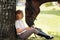 Girl teenager jockey sits in a green clearing under a tree. Feeds a horse an apple and strokes it.