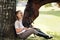 Girl teenager jockey sits in a green clearing under a tree. Feeds a horse an apple and strokes it.