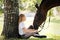 Girl teenager jockey sits in a green clearing under a tree. Feeds a horse an apple and strokes it.