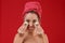 A girl with towel on head puts on face slices of cucumbers. The concept of beauty. On a red background.