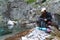 Girl traveler in white hat and black jacket sitting near the mountain lake and opens the backpack