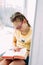 Girl tween with glasses and pigtails reading book at window sill in her room