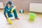 Girl in uniform sitting in squad position and washing floor with hands. She has scrub sponge there. Bottles with liquids