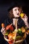 A girl in a vampire costume with yellow eyes holds a basket with vegetables. Bright emotion. Conceptual photo