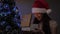 A girl wearing Santa Claus hat opens present next to Christmas tree. Child opens gift box in light of garland, rejoices