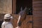 Girl wearing white hat feeds brown horse with flowers woven into mares mane. Horse looks out from behind stable gate and