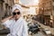 Girl in a white bathrobe and a towel on her head in sunglasses stands on the balcony against the background of the city