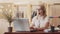 A girl in a white blouse, glasses and headset is sitting at a table with a laptop and typing and talking to customer.