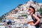 A girl in a white dress, sunglasses and a hat stands on the beach in Positano. View of houses and hotels in the background. Travel