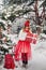 A girl in a white jacket and a red one with Christmas gifts in her hands stands in a snow-covered forest near a Christmas tree.