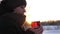 Girl in winter clothes holding in her hand transparent glass with hot tea in winter park at sunset. girl warms her hands
