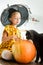 Girl in witch halloween costume sitting on a table playing with pumpkin and her pet cat. Halloween lifestyle concept.