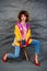 A girl in a yellow jacket and blue jeans with an afro hairstyle sits on a chair. Fashion eighties, the era of disco.