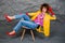 A girl in a yellow jacket and blue jeans with an afro hairstyle sits on a chair. Fashion eighties, the era of disco.