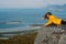 Girl in yellow jacket lies on stone in on the edge of rocks in mountains and looking to fjord