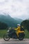 A girl in a yellow raincoat, shoe covers and a white helmet. Motorcyclism and travel. Vertical photo. Top of the Mountains. A gray