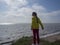 A girl in a yellow windbreaker and red jeans stands on a high beach, looks into the distance, breathes clean air without a mask