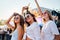 Girls in sunglasses enjoy music at sunny beach fest. Friends dance, laugh with stage behind. Casual summer wear, youth