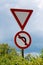 Give way and no left turn red and white signs mounted on strong metal pole surrounded with dense leaves on cloudy stormy blue sky