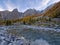 Glacial river with stones in alpine larch forest in autumn, surrounded by mountains