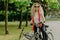 Glad active curly haired female bicyclist covers distance on her own transport, wears sunglasses, casual outfit, poses in park