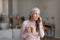 Glad calm caucasian old gray-haired lady with cup of drink speaks by phone in living room interior