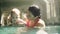 Glad caucasian mother and her baby boy are swimming in the indoors swimming pool, waving him under the water. Child