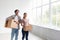 Glad caucasian young guy and woman in casual carry cardboard boxes in empty room interior