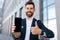 Glad confident young caucasian male with beard in suit shows smartphone with empty screen, shows thumb up