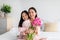 Glad happy japanese teenage girl hugging young woman with bouquet of tulips sitting on bed in bedroom interior
