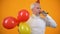 Glad male pensioner using party blower and holding colorful balloons, birthday
