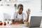 Glad millennial african american female in apron waving hand at laptop in kitchen interior