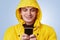 Glad positive male wears yellow anorak and hood, types messages on smart phone, uses free internet connection. Handsome man uses m