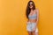 Glad shapely dark-haired girl in black sunglasses standing on yellow background. Indoor photo of slim lady in denim