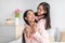 Glad smiling pretty cute asian teenage girl hugging millennial woman in pink pajamas in bedroom interior