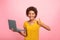 Glad young african american curly female in yellow t-shirt with laptop show thumb up isolated on pink background