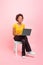 Glad young african american curly lady blogger sit on chair, typing on laptop, isolated on pink background