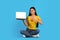 Glad young asian female digital nomad sitting on floor pointing finger at laptop with empty screen