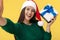 Gladful young brunette woman wear santa claus hat and green pullover take selfie with gift box over yellow background.