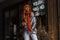 Glamorous young woman in a youth shiny jacket in white trousers in a warm knitted hat with a scarf posing in the city near a