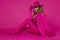 Glamour female model is wearing an elegant fuchsia suit, pink boots and a pink hat and is posing against a pink background.