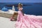 Glamour, stylish elegant bride woman in pink long wedding dress is posing near Church of the Three Bells in Fira in Santorini