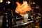 Glass of alcoholic drink and ice stands on bar counter. Bartender makes fire flame over it.