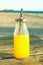 Glass Bottle with Freshly Pressed Tropical Fruits Juice Straw Standing on Plank Wood Table at Beach. Blue Sky Golden Sunlight