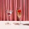 A glass of cocktail and a glass of champagne in front of reach pink renaissance velvet curtain. Dancing glasses retro concept