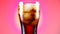 Glass of cola turns slowly around its axis. Close up 4K video. Pink background.