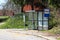 Glass covered transparent local bus station with wooden bench next to traffic signs and notice board surrounded with uncut grass