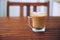 Glass or cup of coffee with white foam on brown wooden table on the balcony, with wooden chair in the background, with copy space